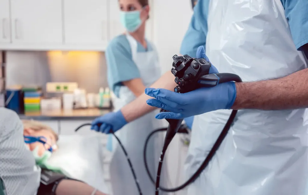 Zdravnik opravlja gastroskopijo pri pacientu, medtem ko drži gastroskop v rokah, oblečen v zaščitna oblačila in rokavice.